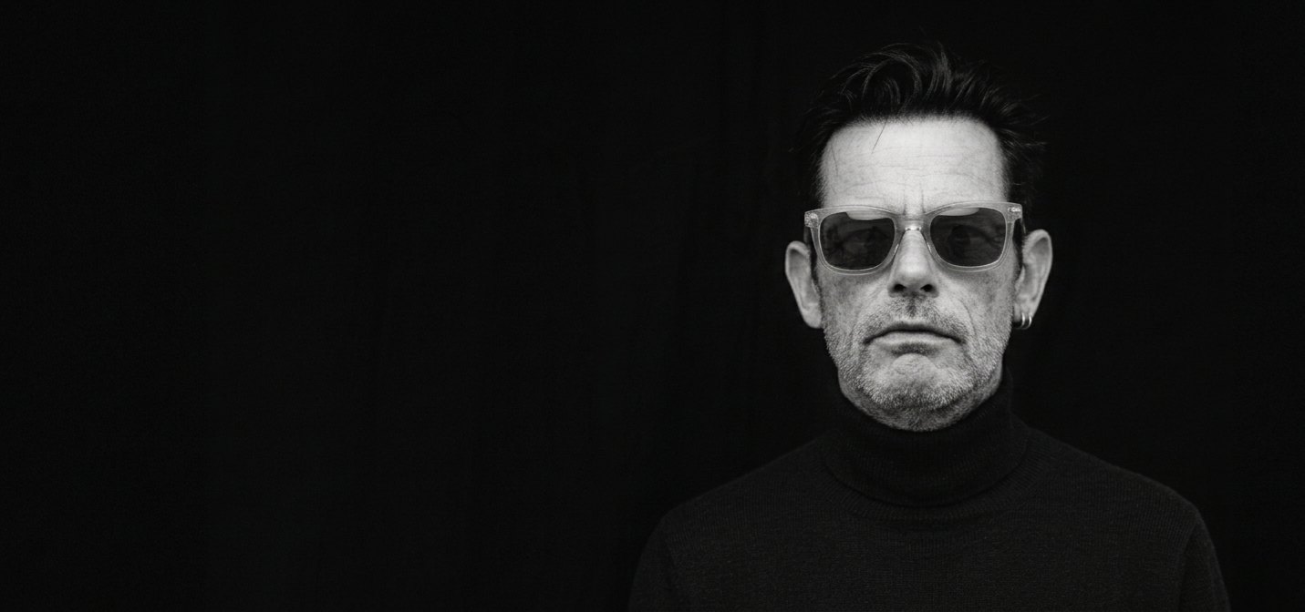 Black and white studio portrait of Hanley in clear-framed sunglasses and a black turtleneck, on a black ground.